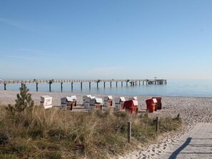 Una playa con un muelle que se adentra en aguas tranquilas, cabañas de playa y un sendero arenoso.