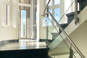 Una escalera moderna con barandillas de vidrio y piso negro, que conduce a un espacio abierto con ventanales grandes.