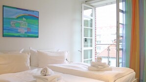 A hotel room with two beds, white bedding, a painting on the wall, and a view of buildings through an open door.