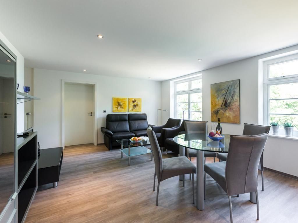 Una sala de estar moderna con una mesa de comedor de vidrio, sillas grises, un sofá negro y una mesa de centro. Hay dos cuadros en la pared y una ventana con vistas a zonas verdes.