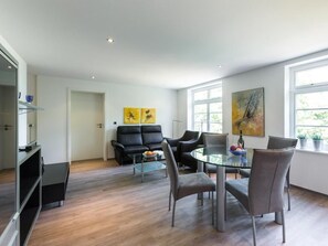 A modern living room with a glass dining table, grey chairs, a black sofa, and a coffee table. There are two paintings on the wall and a window with a view of greenery. - Villa Höger Whg. 09, Deutschland (Ostseebad Boltenhagen)