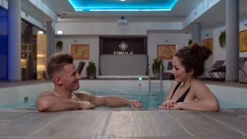 Una pareja sentada junto a una piscina en un ambiente interior moderno con un logotipo visible en la pared.