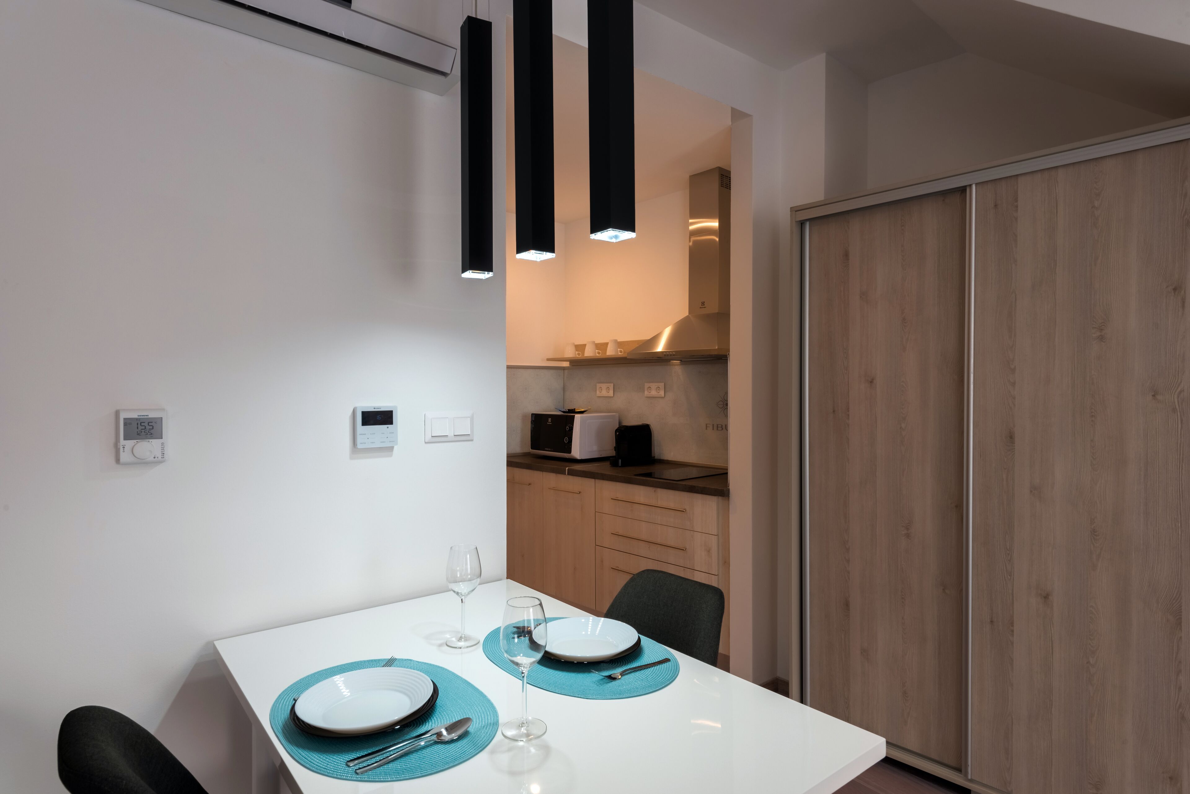 A modern dining area with a white table, teal placemats, and black chairs. A kitchen is visible in the background with wooden cabinets and a microwave.