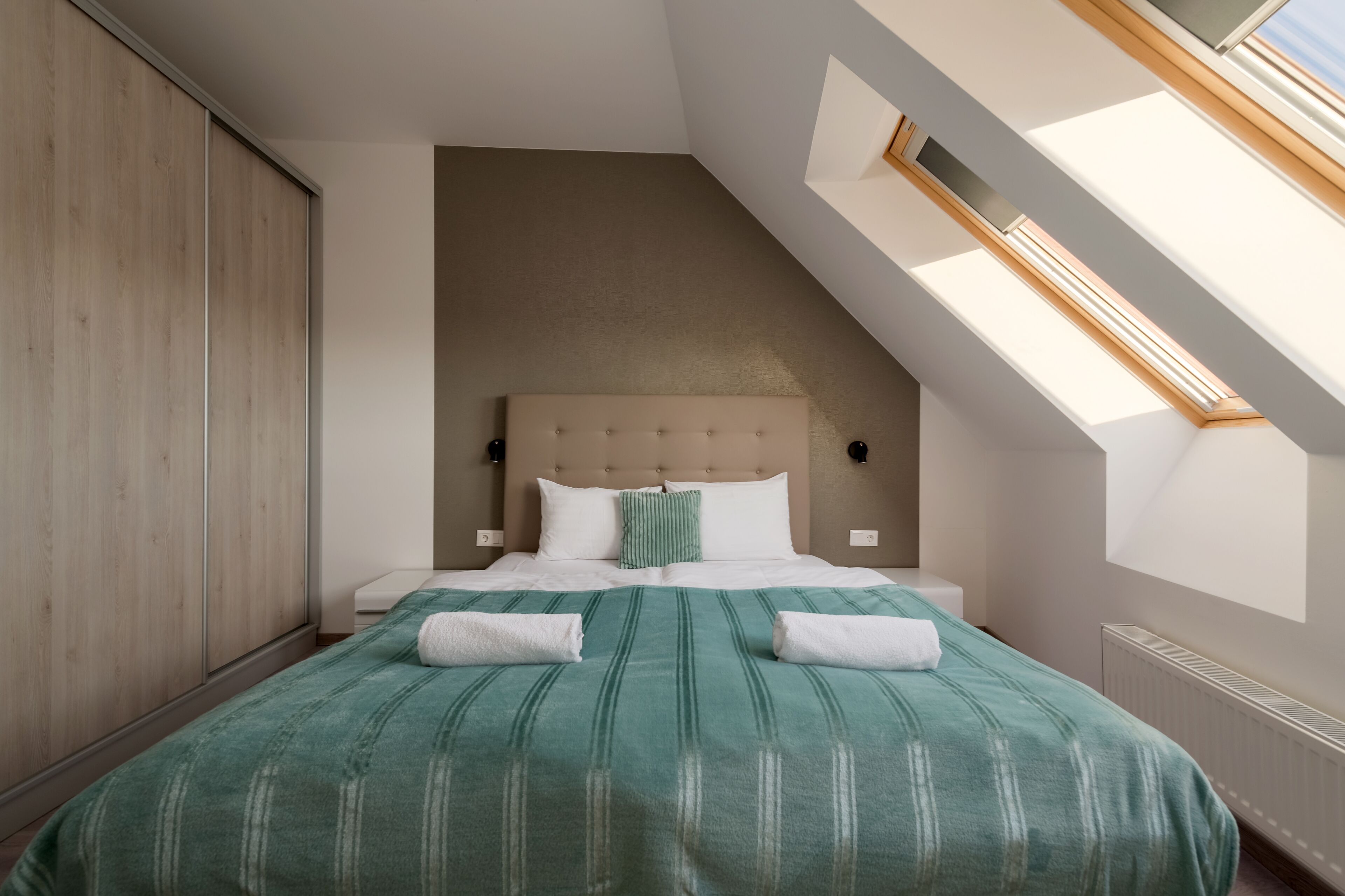 Ein Schlafzimmer mit schrägem Dach, einem Bett mit grüner Bettwäsche, einem Holzschrank und einem Oberlicht.