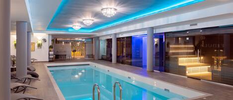 A modern indoor swimming pool with a clear water surface, surrounded by lounge chairs and a reception area with a marble wall.