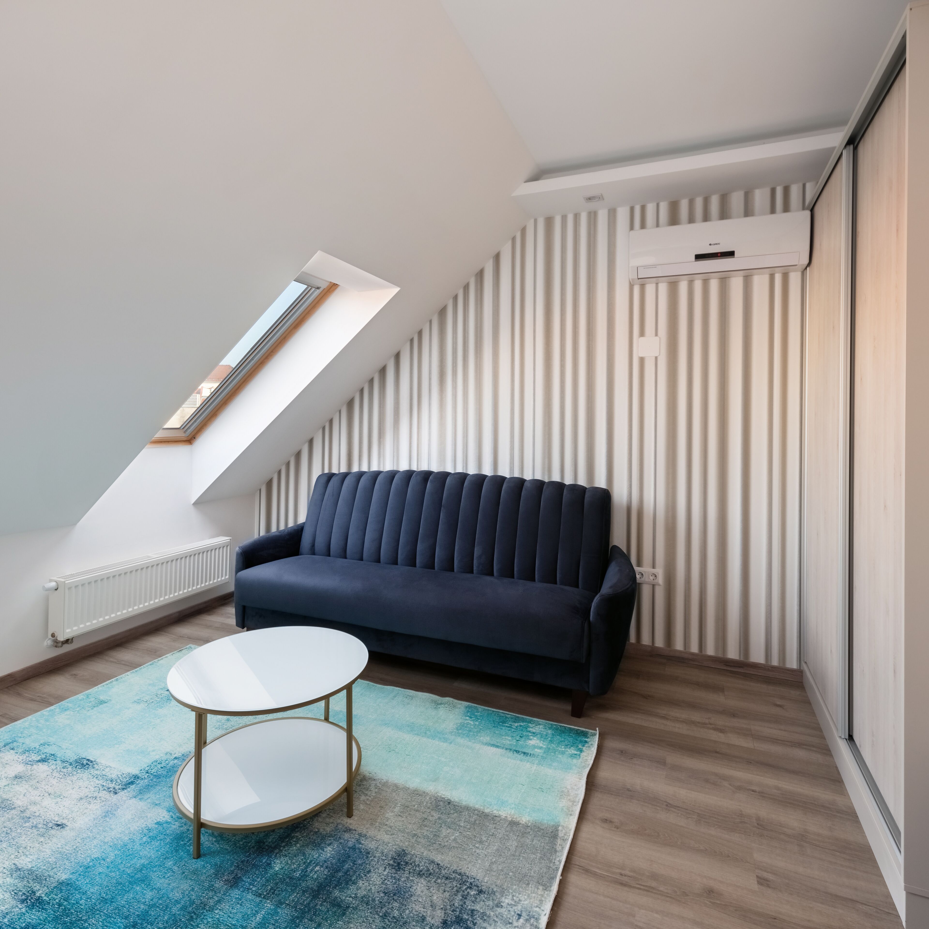 Une chambre à coucher de style loft moderne, avec un plafond en pente, un canapé bleu foncé, une table basse ronde blanche et un tapis à motifs bleus et blancs.