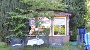 Una pequeña construcción de madera con una ventana, rodeada de follaje y sillas metálicas con cojines.
