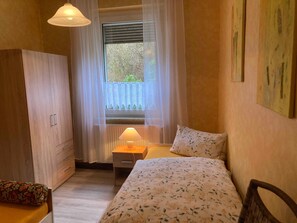 Une chambre à coucher avec un lit, une table de chevet, une lampe, une armoire, une fenêtre avec des rideaux et un tableau au mur.