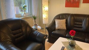 Una sala de estar con dos sofás de cuero negro, una mesa de centro blanca, un control remoto y un florero con una rosa roja.