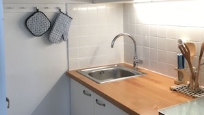 Une cuisine avec des armoires blanches, un évier en acier inoxydable et un plan de travail en bois. Deux maniques à motifs sont accrochées au mur.