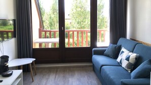 Une pièce avec un canapé bleu, une petite table sur laquelle se trouve une machine à café, et un téléviseur fixé au mur. Deux portes-fenêtres coulissantes donnent sur un balcon extérieur doté d’une rambarde rouge.