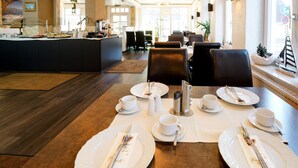 A dining area with tables set for four, each with white plates, cups, and cutlery. There are salt and pepper shakers, a coffee machine, and a buffet counter in the background. - Double room - Hotel Zur Mühle Ziegler family (Kappeln)