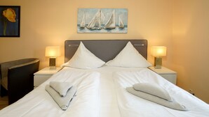 Una cama bien hecha con sábanas blancas, dos almohadas y toallas dobladas. Hay dos mesitas de noche con lámparas, un cuadro de veleros en la pared y una silla a la izquierda.
