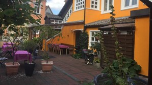 Un patio con mesas al aire libre, plantas en macetas y un edificio amarillo con marcos de ventana blancos.