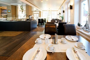 A dining area with tables set for four, each with white plates, cups, and cutlery. There are salt and pepper shakers, a coffee machine, and a buffet counter in the background.