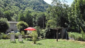 Uma área externa gramada com uma mesa, cadeiras, um guarda-sol vermelho e uma estrutura de madeira rústica.