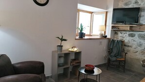 Uma sala de estar com um sofá, uma mesa pequena com uma tigela, uma planta em vaso, um relógio de parede, uma televisão e uma parede de pedra.