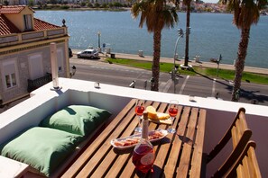 A balcony with a wooden table, green cushions, a bottle of wine, and a plate of food. - Loft with terrace and river view in Seixal. Air conditioning. Ideal for 2pax. (Seixal)