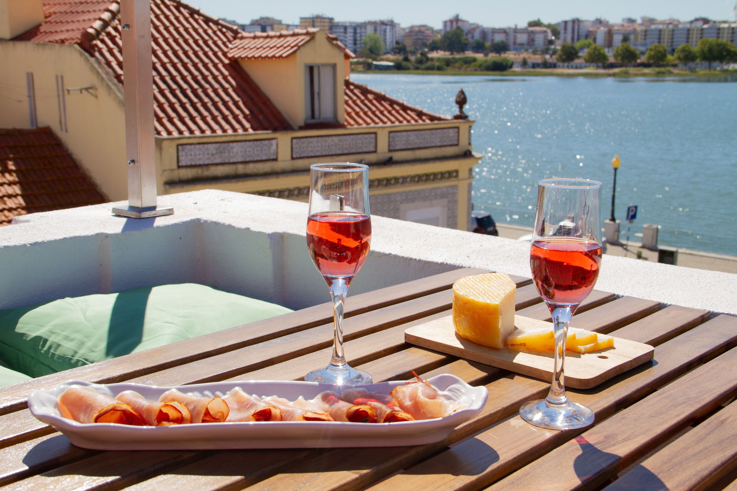 A dining table with a plate of cured meats, two glasses of red wine, and a piece of cheese on a wooden deck overlooking a body of water with buildings in the background.