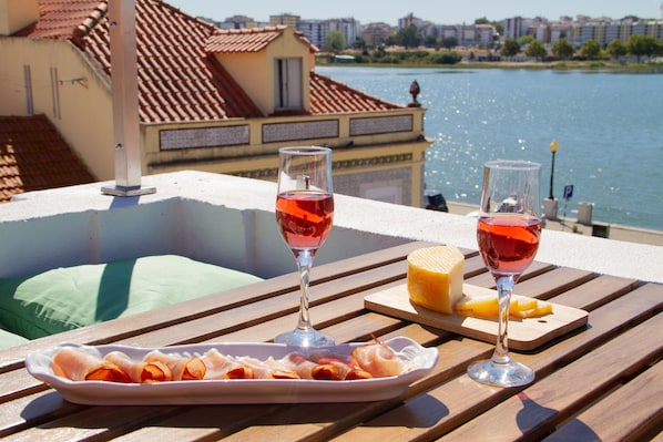 A dining table with a plate of cured meats, two glasses of red wine, and a piece of cheese on a wooden deck overlooking a body of water with buildings in the background.