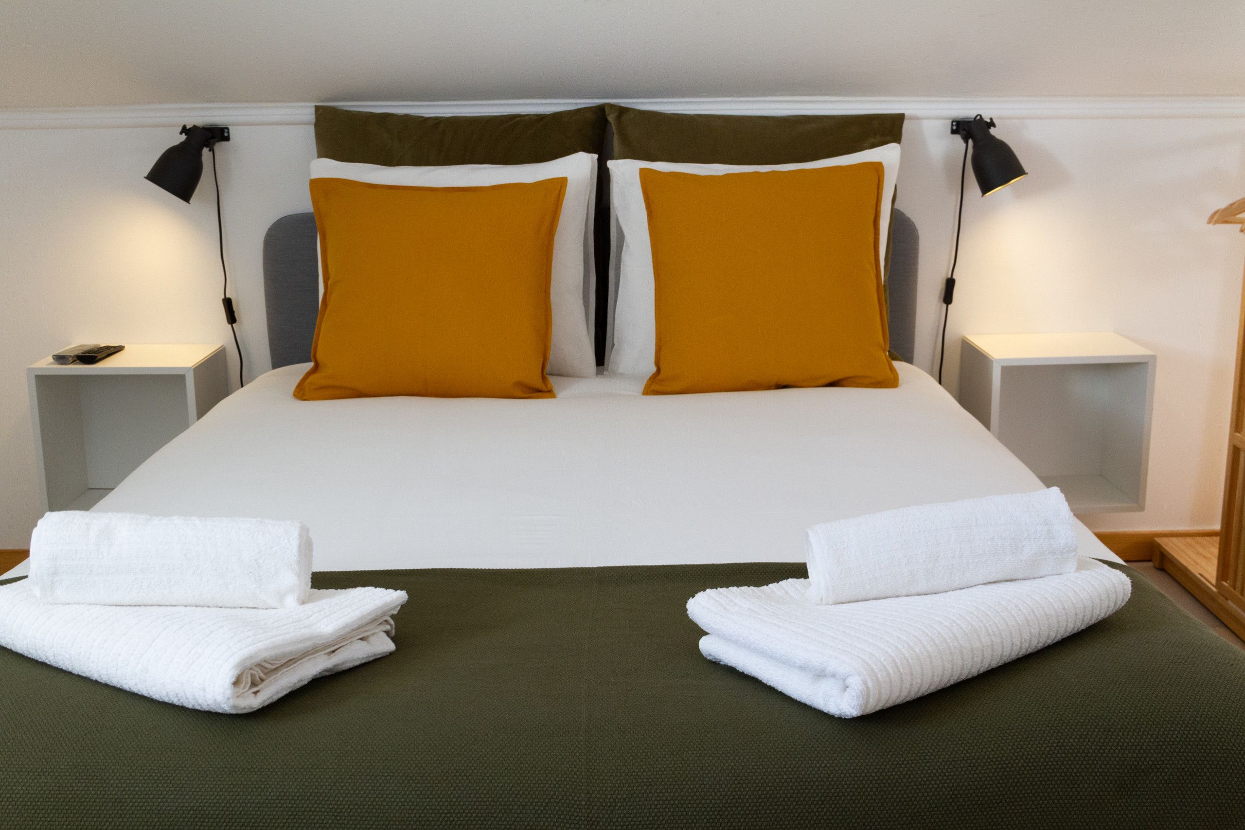 Cama bem arrumada com duas toalhas brancas dobradas no pé, um edredom verde e dois travesseiros amarelo-mostarda.