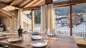 Un coin repas avec une table en bois dressée pour le repas, de grandes fenêtres avec des rideaux et une vue sur un paysage enneigé avec des bâtiments.