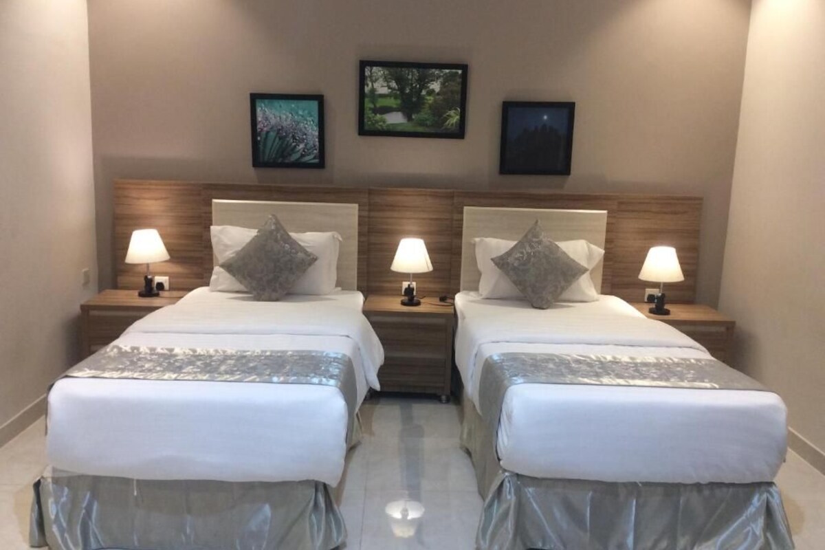 A hotel room with two beds, each with a wooden headboard, white bedding, and a grey skirt. There are bedside lamps and framed pictures on the wall.