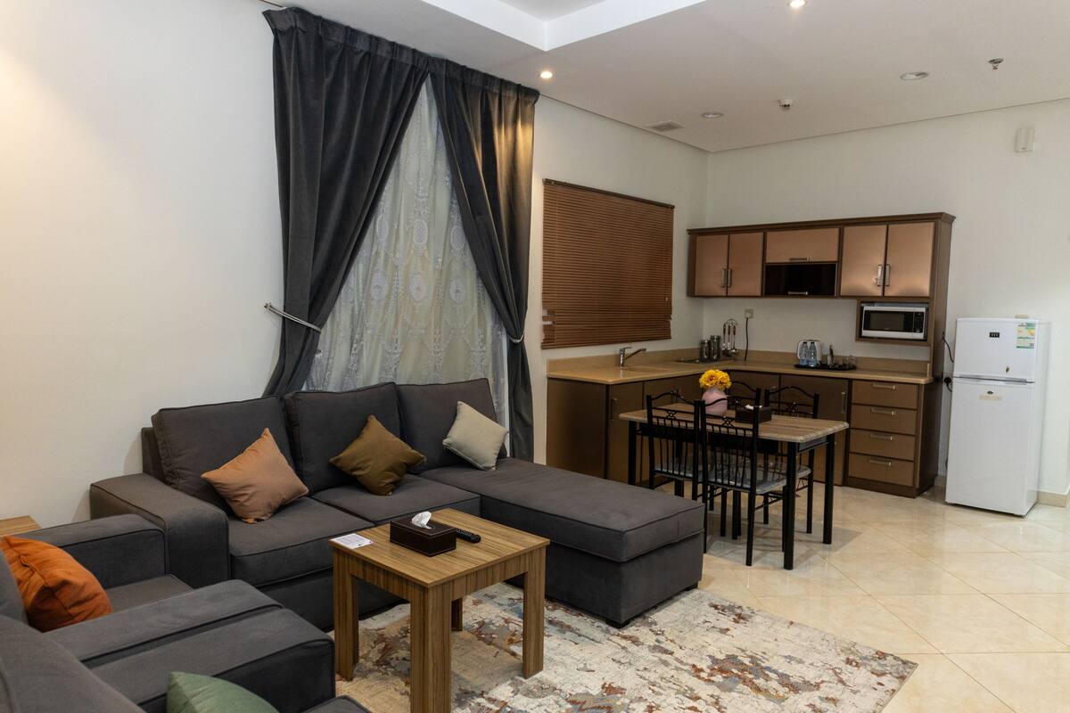 A modern living room with a grey sectional sofa, a wooden coffee table, and a patterned rug. Adjacent to the living room is a compact kitchen with a microwave, refrigerator, and wooden cabinets.