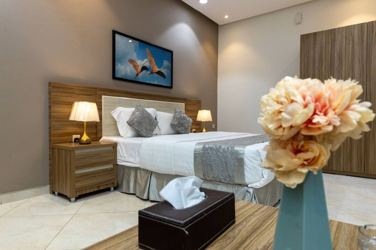 A modern bedroom with a large bed, wooden headboard, bedside lamps, a nightstand, and a vase with flowers.