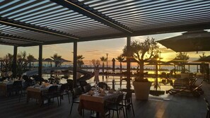 Uma área de refeições ao ar livre com mesas e cadeiras, piscina e vista para o pôr do sol.