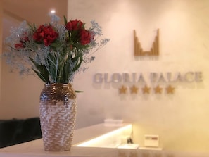 Een vaas met rode bloemen en ademtak op een aanrecht, met een bordje met de tekst 'GLORIA PALACE' en drie sterren.