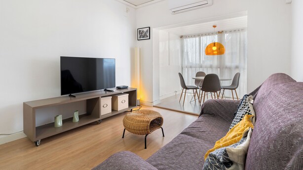 Een moderne woonkamer met een flatscreen televisie, een bank, een rieten voetenbankje en een eethoek met stoelen en een tafel.