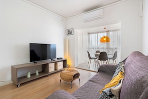 A modern living room with a flat-screen TV, a sofa, a wicker ottoman, and a dining area with chairs and a table.