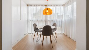 Een kamer met een ronde tafel, vier stoelen, een hanglamp en grote ramen met dunne gordijnen.