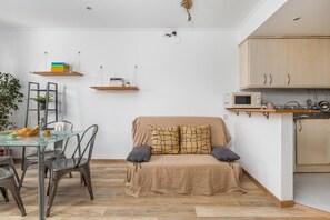A compact living space with a glass dining table, metal chairs, a sofa with patterned cushions, a kitchen area with wooden cabinets, and wall-mounted shelves.