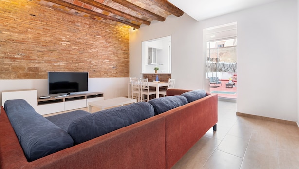 A modern living room with a brick wall, a flat-screen TV, a sofa, a dining area with chairs, and a large window.