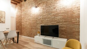 A modern living room with a brick wall, a flat-screen TV on a white cabinet, and a dining area with chairs and a table. - Unpack your bags, take a deep salty breath, inhale the sea in, and wonder at the magic of Barcelona (Barcelona)