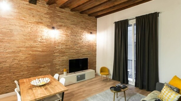 A cozy living room with a brick wall, wooden ceiling beams, a flat-screen TV, a wooden dining table, and a sofa with a yellow cushion. - Unpack your bags, take a deep salty breath, inhale the sea in, and wonder at the magic of Barcelona (Barcelona)