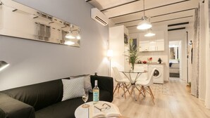 Un salon moderne avec un canapé noir, une petite table ronde sur laquelle se trouvent un livre et des lunettes, ainsi qu’une bouteille d’alcool. Un verre de vin est posé sur la table. La pièce est dotée d’un grand miroir au mur et d’un plafond avec des poutres apparentes. On aperçoit un coin cuisine au fond de la pièce.