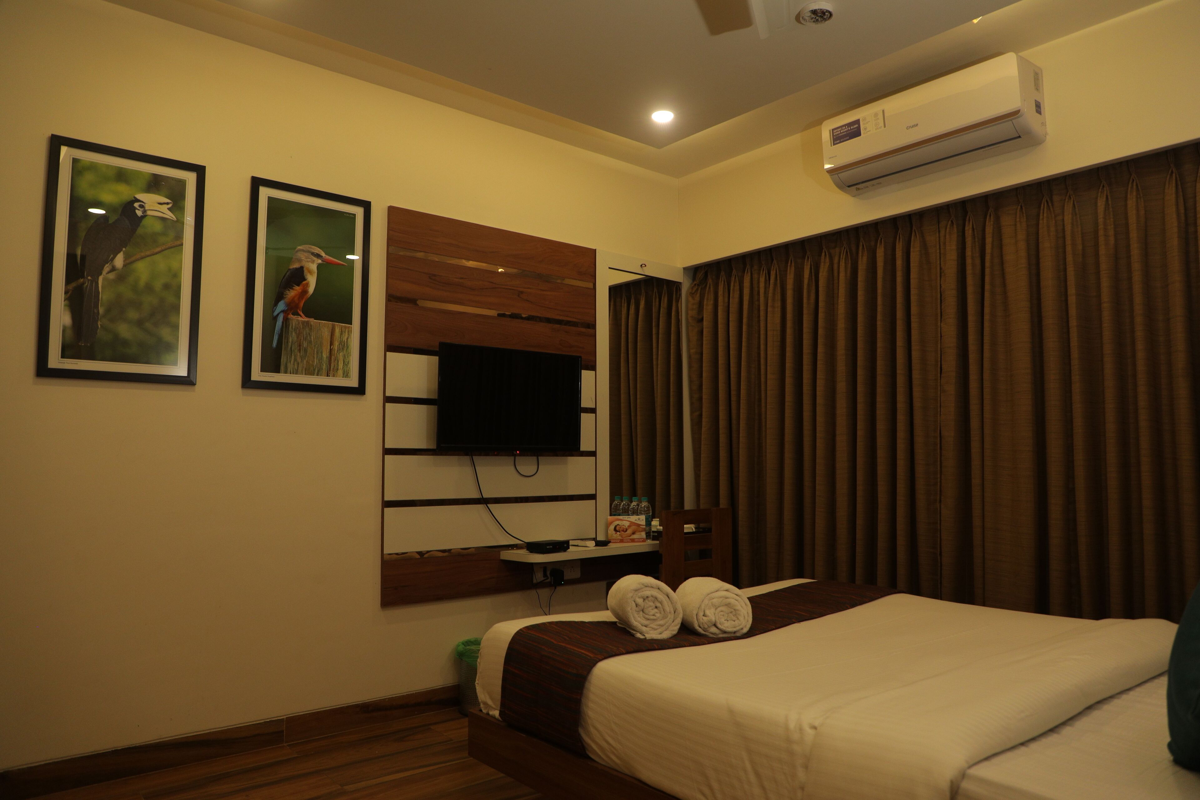 A hotel room with a bed, a flat-screen TV mounted on the wall, a wooden headboard, and a window with curtains.