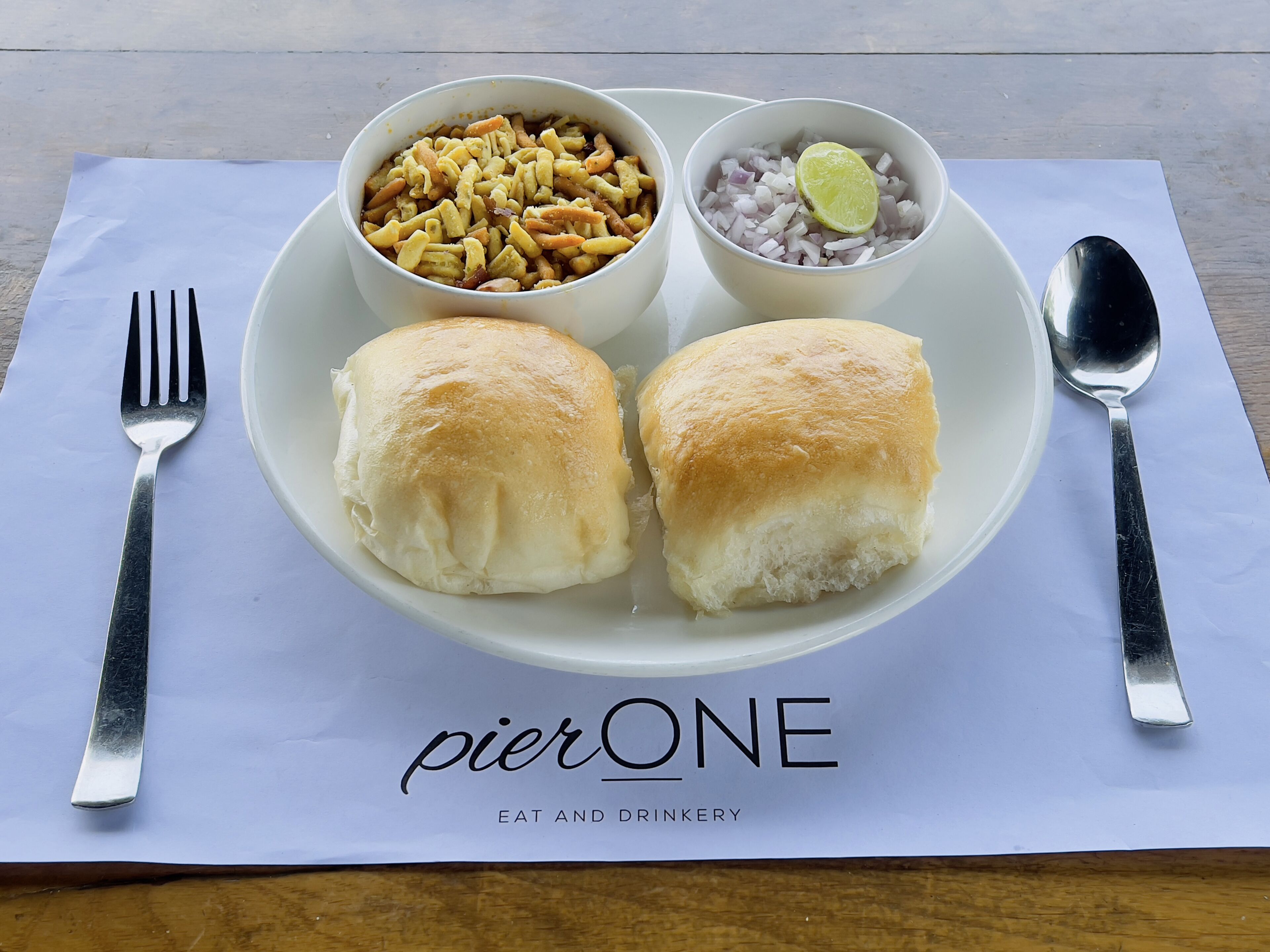 A plate with two rolls of bread, a bowl of nuts, and a bowl of diced onions with a lime slice on a white napkin with "pierONE" branding.