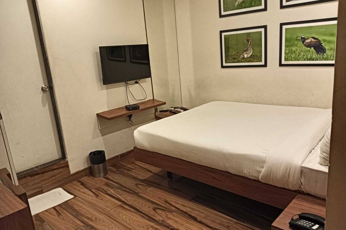 Um quarto com uma cama, uma televisão presa à parede, uma pequena mesa de cabeceira de madeira, um cesto de lixo e quadros pendurados na parede.