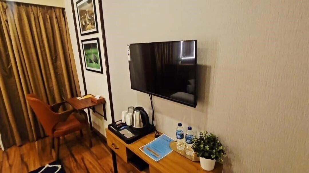 A hotel room with a flat-screen TV mounted on the wall, a wooden desk with a kettle, a chair, a small table with water bottles, and a potted plant.