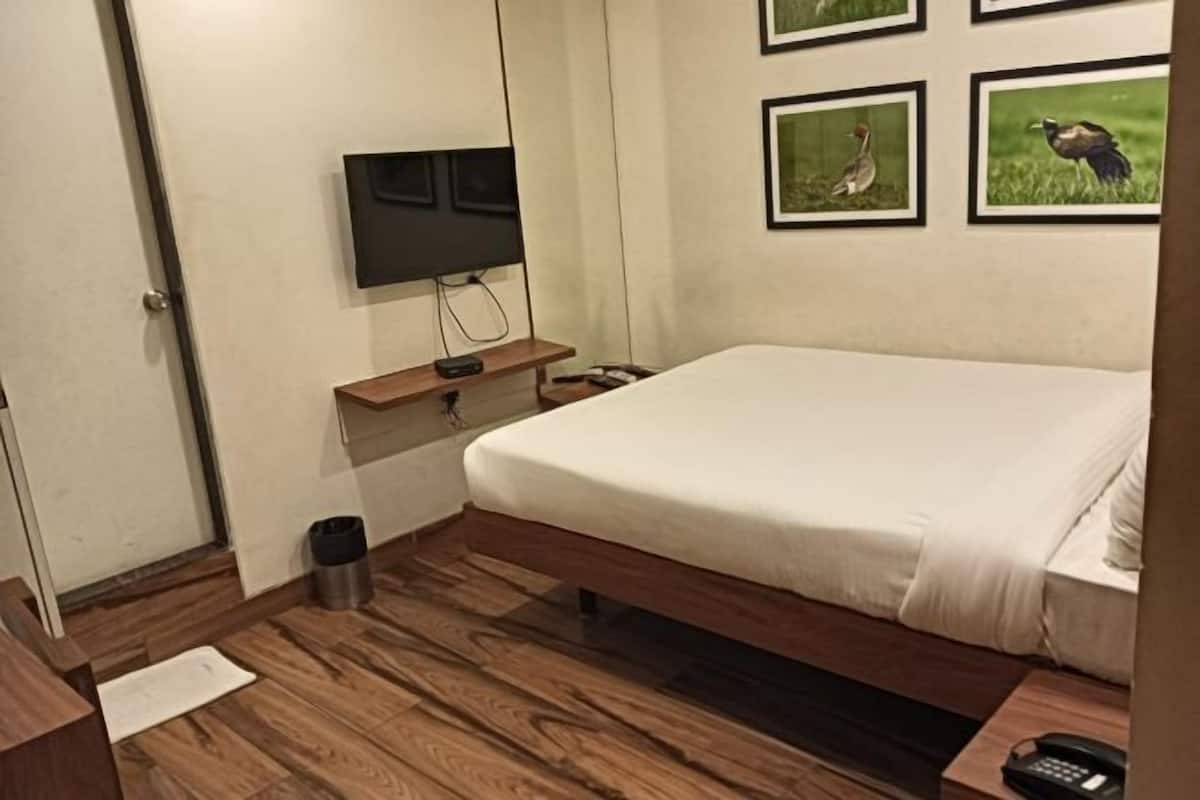 Quarto de hotel com uma cama, uma televisão presa na parede, uma mesinha de cabeceira de madeira e quadros na parede.