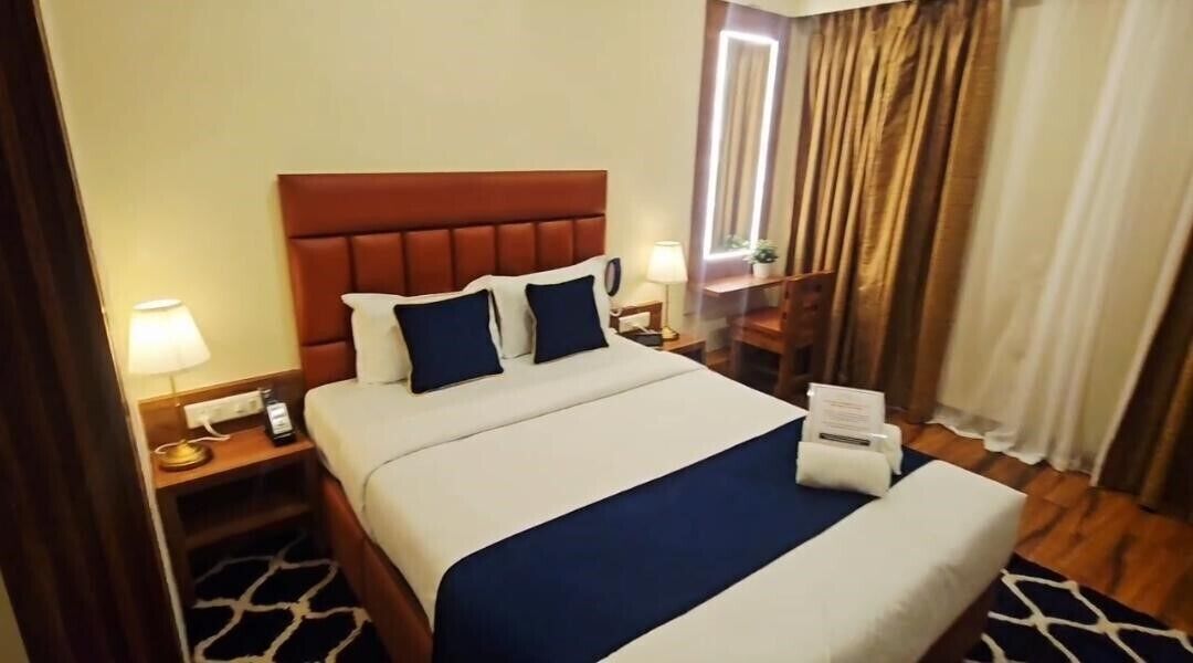 A hotel room with a large bed, a nightstand with a lamp, a bedside table with a phone, and a window with curtains.