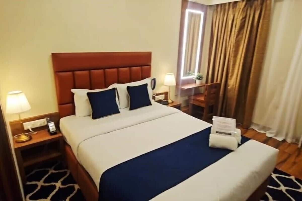 Quarto de hotel com uma cama grande, mesa de cabeceira com abajur, criado-mudo com telefone e janela com cortinas.