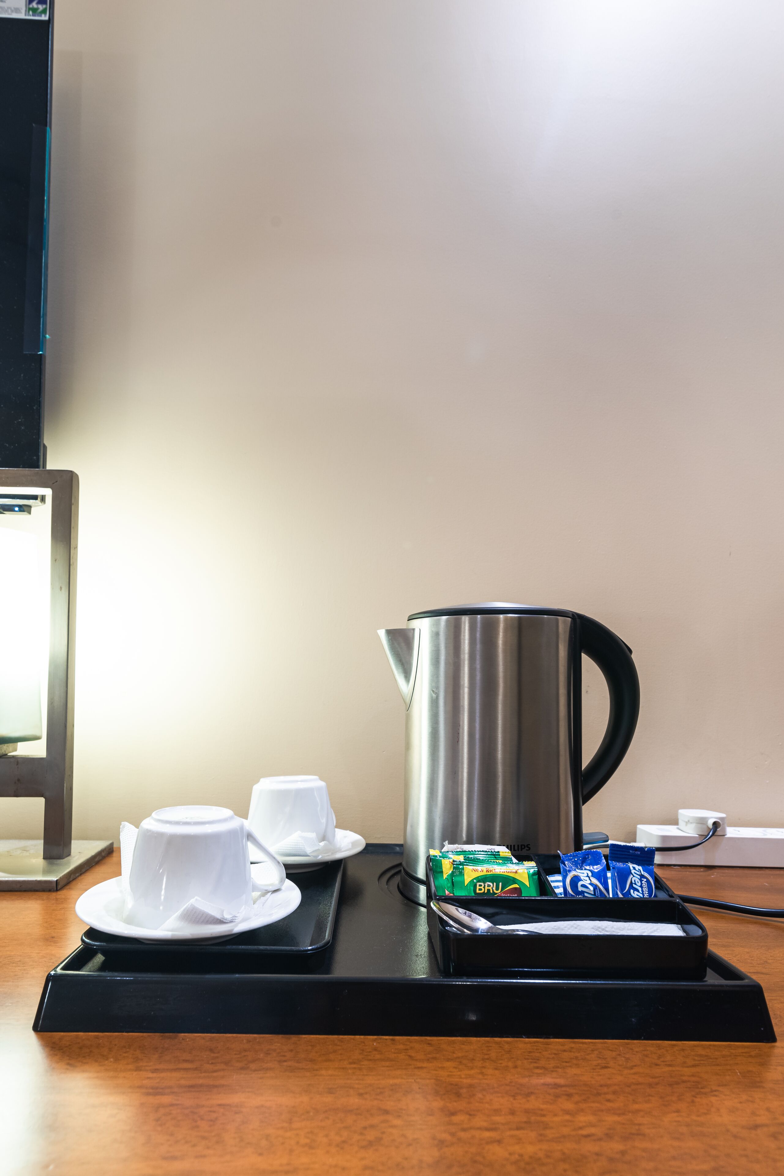 Una bandeja con un hervidor de acero inoxidable, dos tazas blancas y algunos alimentos envasados sobre una mesa de madera.
