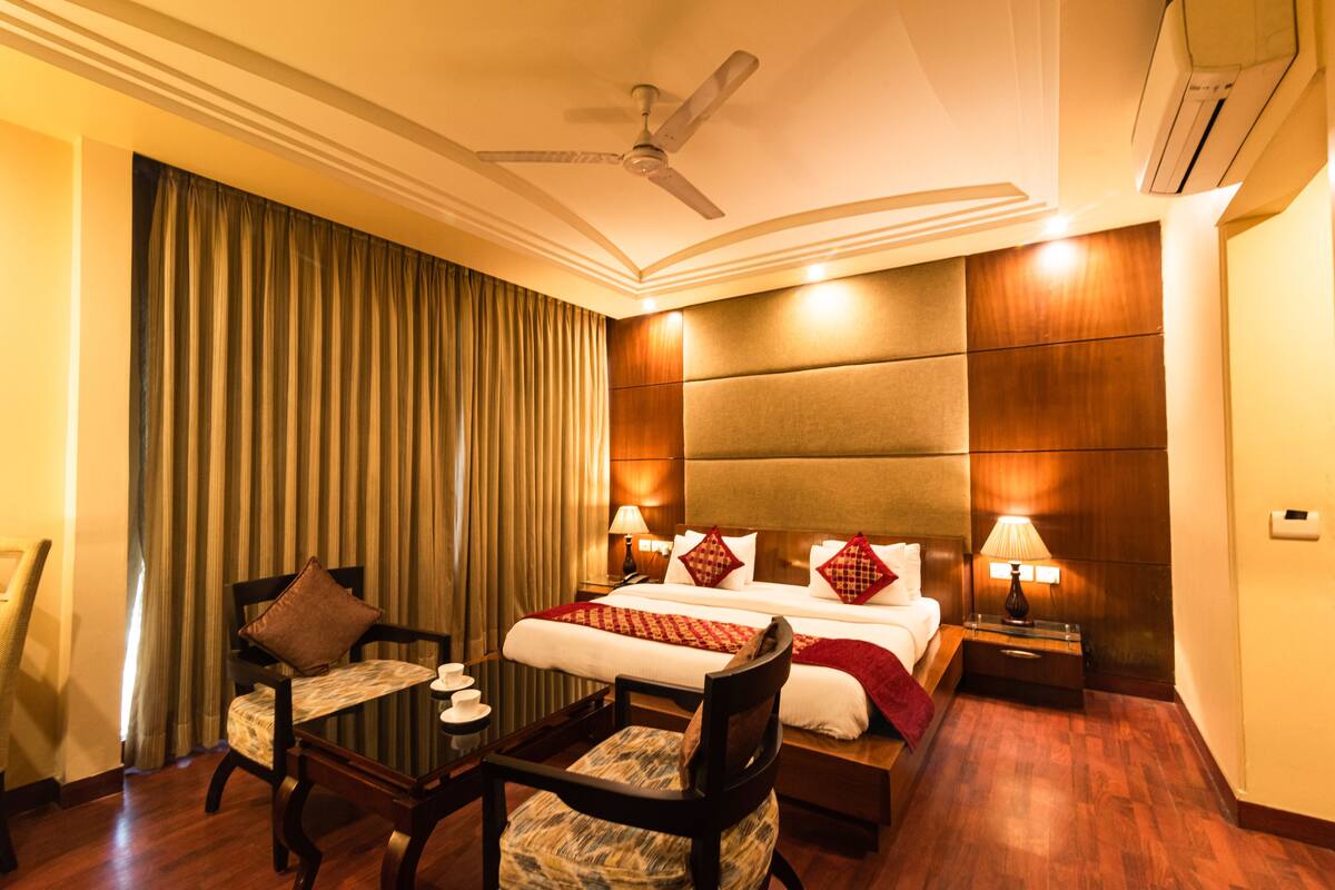 A hotel room with a bed, two bedside tables with lamps, a chair, a desk, a ceiling fan, and a large window with curtains.