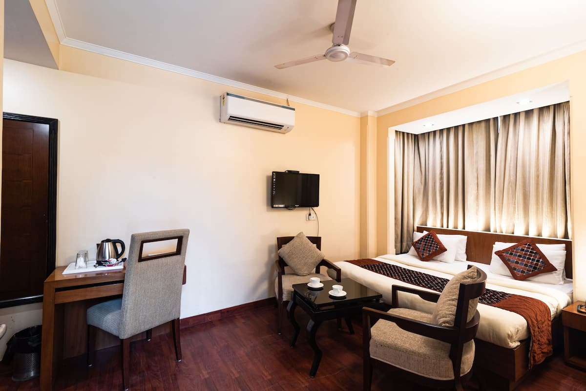 A hotel room with a bed, desk, chairs, a television, and an air conditioning unit.