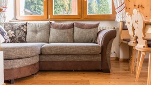 A corner sofa with a view of a forest through a window, a wooden chair, and a radiator.